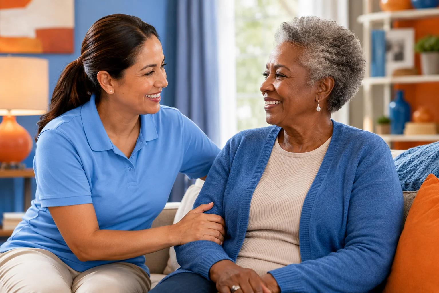 A smiling Hispanic caregiver provides companionship and support to an elderly African American woman in a comfortable home setting.